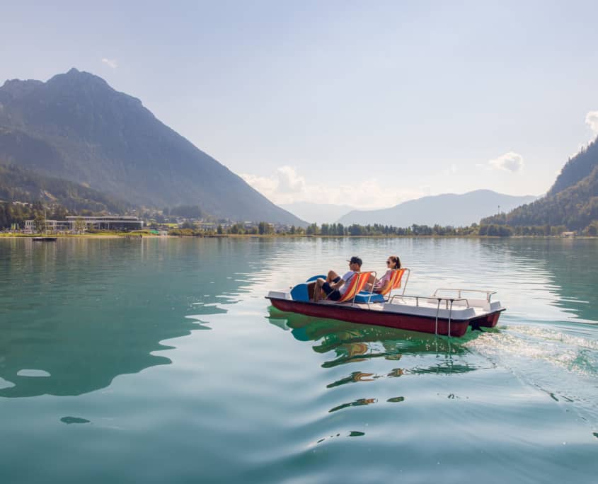 Zwei Menschen genießen das Schwimmen im Achensee auf einem Boot mit Bergen im Hintergrund.